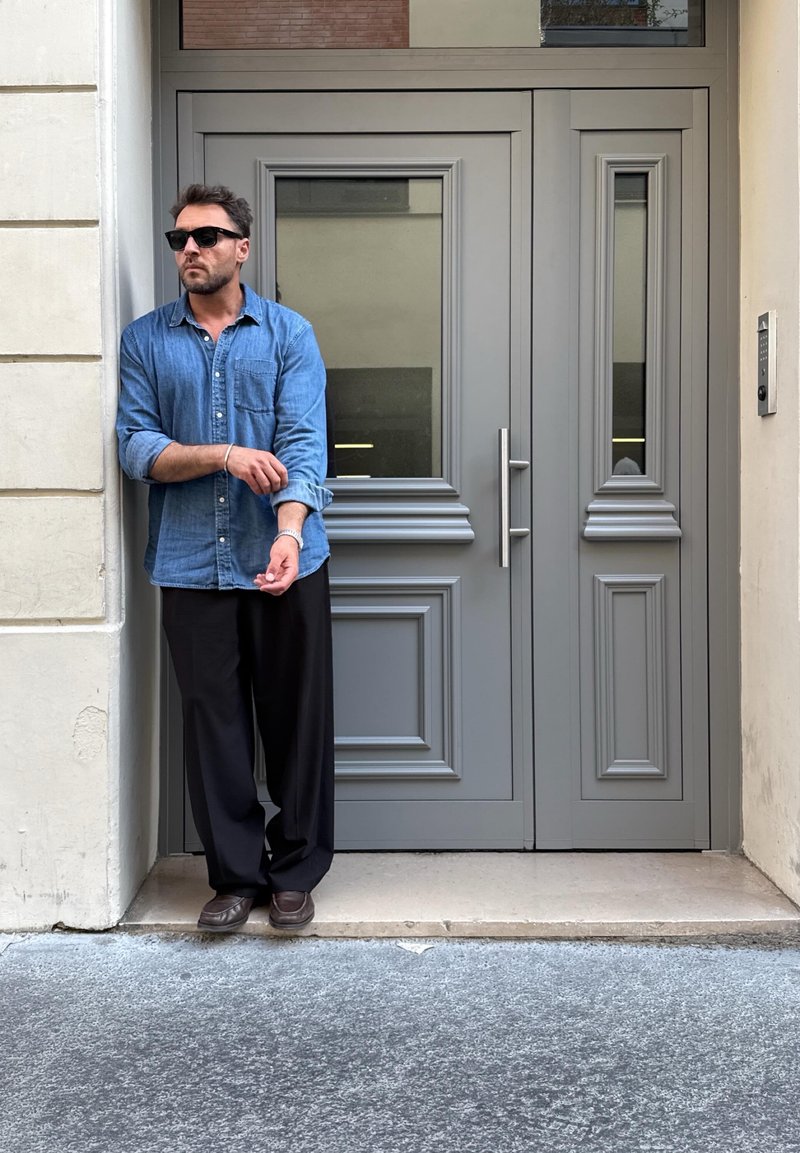 Homme portant des lunettes de soleil, une chemise bleue et un pantalon noir, debout, retroussant sa manche, à côté de grandes portes doubles grises dans une rue urbaine.