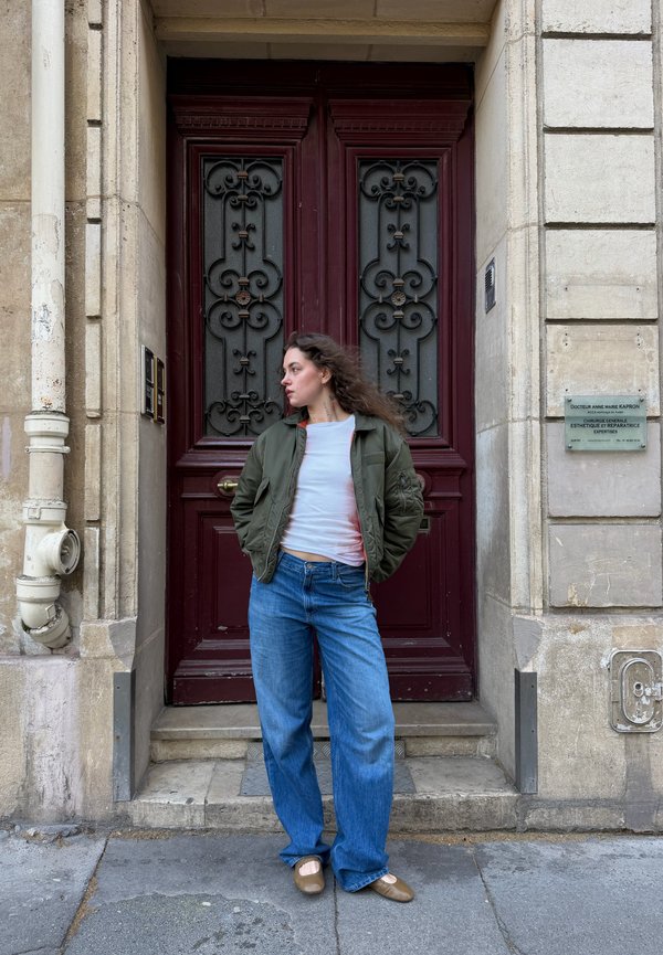 Junge Frau mit langen Haaren steht und schaut nach links, trägt eine grüne Jacke, ein weißes Hemd, weite blaue Jeans und braune Schuhe vor einer großen verzierten dunkelroten Tür.