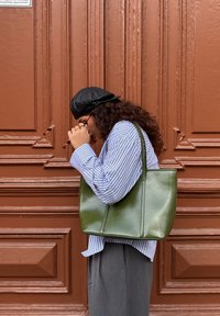Sac cabas vert olive en matière brillante, avec une forme haute, deux anses et des détails de coutures verticales. Porté avec une chemise à rayures.