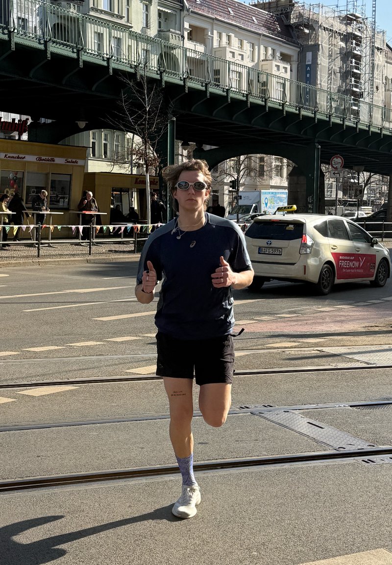 Løber med solbriller og mørkeblå sportstøj jogger på en byvej med sporvognsskinner, taxa og forhøjet jernbane i baggrunden på en solskinsdag.