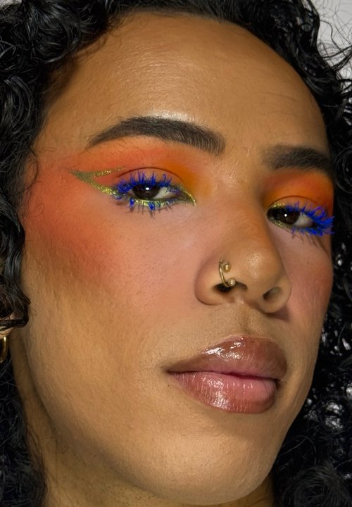 Person mit lockigem Haar, die blaue und silberne Lidschatten mit kleinen Strasssteinen trägt, neutralen Lippenstift und einen goldenen Nasenring.