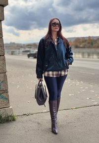 Marineblå blank jakke lagdelt over en ternet skjorte, kombineret med mørke leggings og knælange brune støvler, holdt med en grå håndtaske.