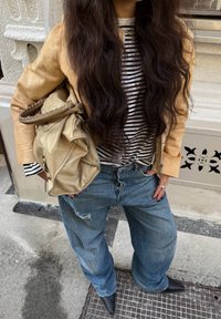 Beige Lederjacke, schwarz-weiß gestreiftes Hemd, zerrissene blaue Jeans und schwarze spitze High Heels. Trägt eine weiche beige Tasche.