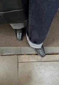 Zapatos de cuero negro con punta afilada y diseño texturizado, combinados con vaqueros oscuros con los bajos remangados, sobre una superficie texturizada.