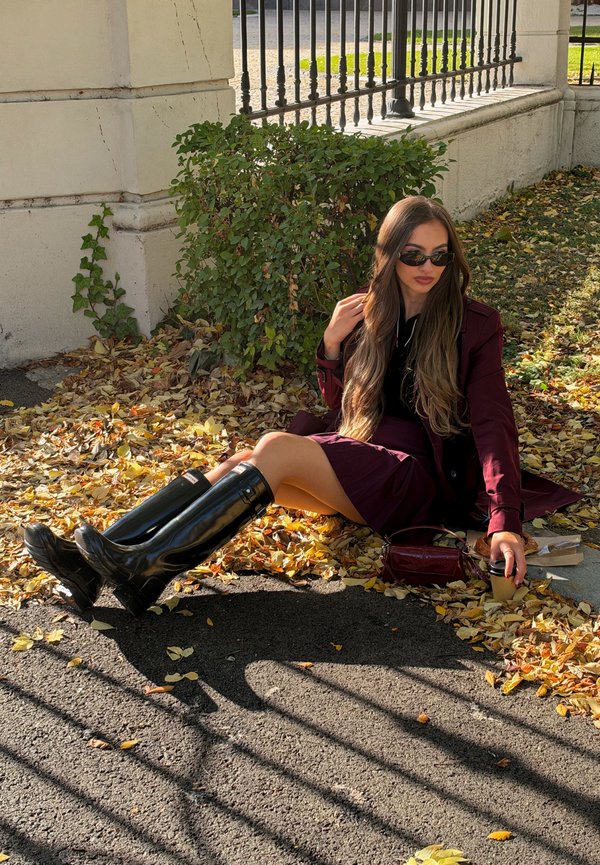 Una mujer con gafas de sol y un conjunto color burdeos está sentada entre hojas otoñales junto a un muro de piedra y sostiene un vaso para llevar.