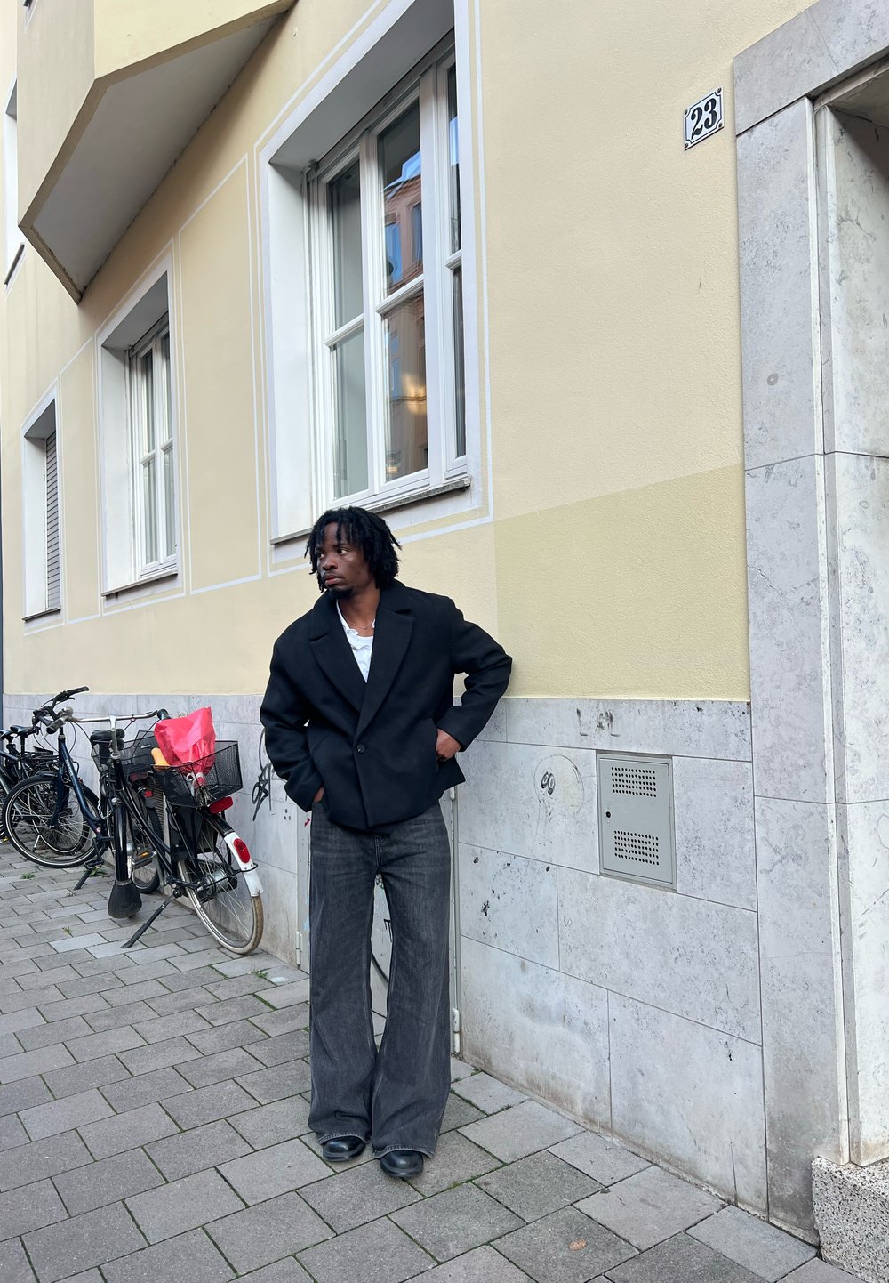 Czarny oversize'owy płaszcz z klapowanym kołnierzem, biała koszula pod spodem oraz szerokie ciemne dżinsy z rozszerzanymi nogawkami; stoi obok ściany, obok rowerów.