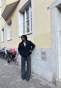 Czarny oversize'owy płaszcz z klapowanym kołnierzem, biała koszula pod spodem oraz szerokie ciemne dżinsy z rozszerzanymi nogawkami; stoi obok ściany, obok rowerów.