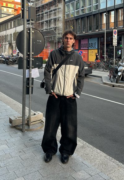 Pantalones de pierna ancha negros, chaqueta con cremallera gris y blanca, zapatos negros, de pie en una acera con elementos de fondo urbano, incluyendo bicicletas y una señal de tráfico.