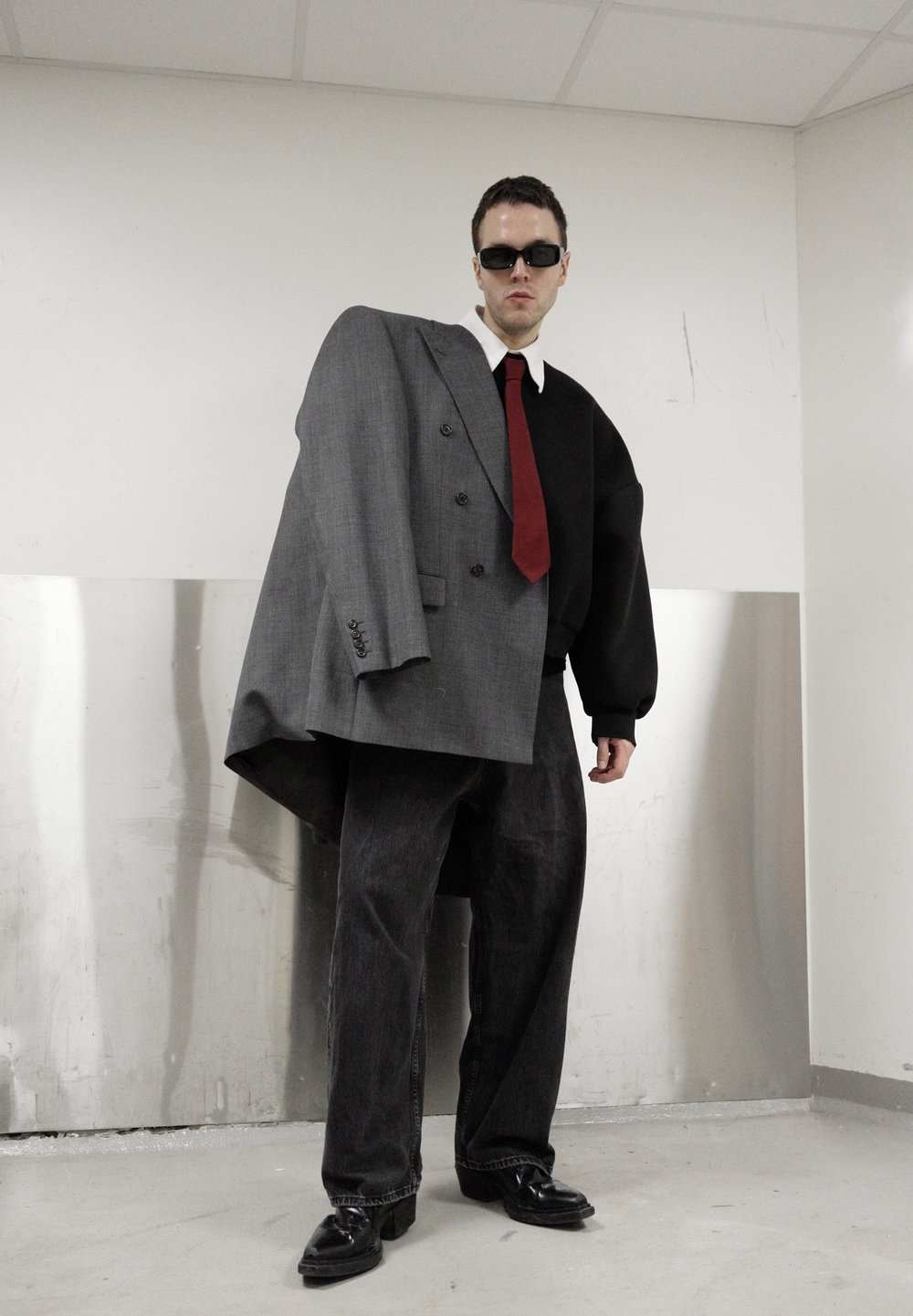 Veste gris surdimensionnée à double boutonnage associée à un pull noir surdimensionné, cravate rouge, jean noir et chaussures noires brillantes. Surface de sol mate.