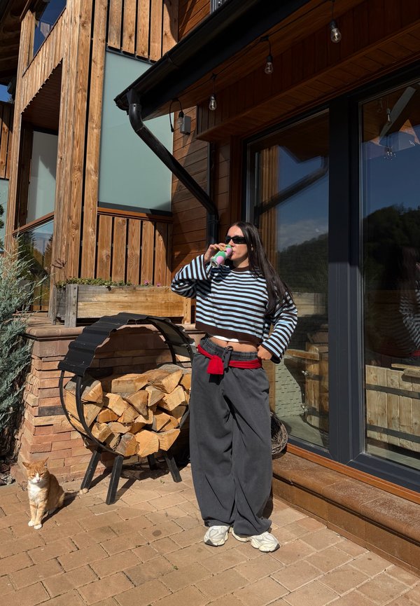 Femme portant des lunettes de soleil et une chemise rayée boit dans une canette devant une maison en bois avec du bois de chauffage empilé et un chat roux à proximité.