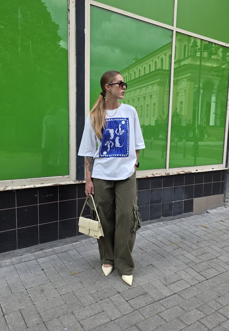 T-shirt blanc surdimensionné avec un design graphique bleu, pantalon cargo vert olive, chaussures jaunes à bout pointu et un petit sac à main crème.