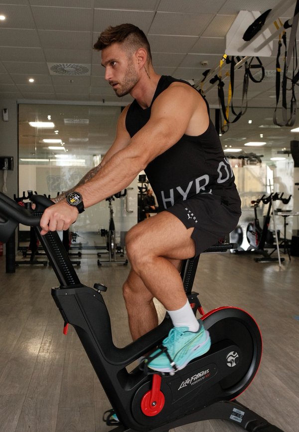 Un hombre con ropa deportiva pedalea en una bicicleta estática en un gimnasio, sujetando el manillar.