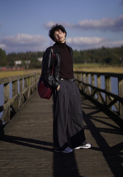 Chaqueta de cuero negra sobre un suéter de cuello alto marrón, pantalones grises de pierna ancha y zapatillas plateadas. Bolso burdeos con un patrón texturizado. Fondo de un muelle de madera.