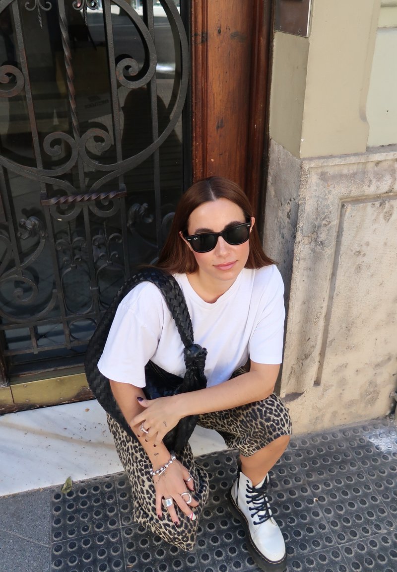 T-shirt surdimensionné blanc, sac à main noir tressé, shorts imprimé léopard, bottes argentées à lacets et lunettes de soleil. Fond en marbre et pavé texturé.