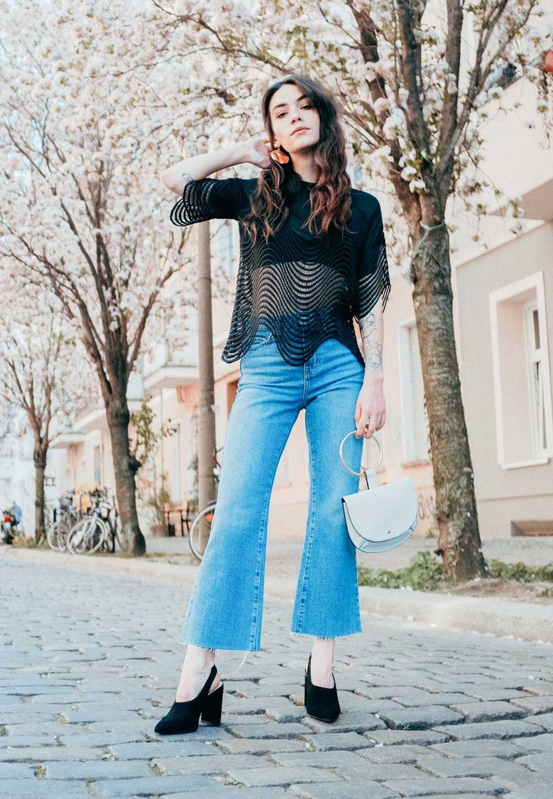 Fekete hálós felső hullámos szegéllyel, világoskék trapéznadrág, fekete magassarkú cipő, egy kis fehér kézitáska a kezében, háttérben cseresznyevirág fákkal.