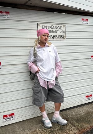 T-shirt blanc surdimensionné par-dessus une chemise à carreaux rose, shorts cargo gris, chaussettes rose clair et baskets grises. Bandana rose, sac argenté.
