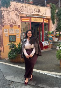 Bordeaux nederdel, beige sweater og grå jakke; står foran en kunstbutik med farverige malerier og teksturerede vægge udsmykket med dekorationer.