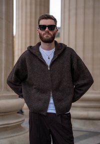 Sweat à capuche marron à fermeture éclair, fabriqué en tissu texturé avec une capuche et un ourlet côtelé, porté par-dessus une chemise blanche, associé à un pantalon noir et des lunettes de soleil.