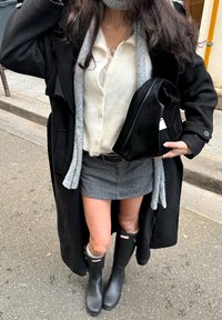 Žena v černém kabátě, bílém kardiganu, šedé sukni, černých kozačkách po kolena a s černou kabelkou na městském chodníku.