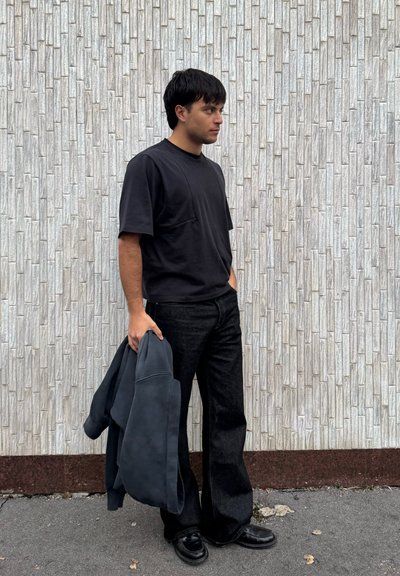 Joven con cabello negro, vistiendo una camiseta negra y pantalones vaqueros anchos negros, sosteniendo una chaqueta oscura, de pie frente a una pared de piedra texturizada.