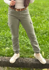 Ljust grå cargo-byxor med elastiska muddar, sidofickor och ett brunt bälte. Bärs med vita sneakers på en grönskande bakgrund.