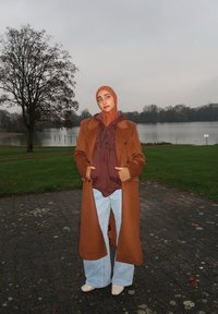 Brun uldfrakke lagdelt over en brun hættetrøje, kombineret med lyseblå vidde jeans og cremefarvede sko. Naturlig baggrund.