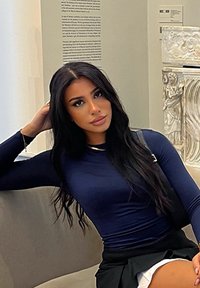 Marinblå långärmad topp med en figurnära design, matchad med en svart kjol. Modellen har långt mörkt hår och sitter på en ljus soffan.