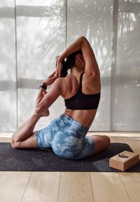 Yoga poza na crnoj prostornoj prostirci. Subjekt nosi crni sportski grudnjak i plave šortseve s batik uzorkom, a pored njega je pluta blok. Svjetlost iz prozora.