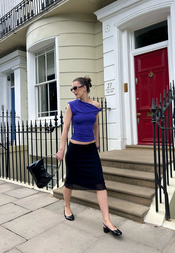 Femme en haut violet sans manches et jupe noire marchant devant une maison avec une porte rouge et une balustrade noire, tenant un sac à main noir dans la main gauche.