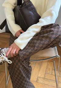 Personne assise sur une chaise en métal, portant un pantalon marron à motifs, un pull crème, des chaussettes beiges, tenant un sac à main en cuir marron foncé sur les genoux.