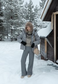 Casaco puffer cinza com capuz forrado de pele, suéter de malha com padrão geométrico, calças cinzas com textura e corte amplo, e bolsa crossbody bege em uma paisagem nevada.