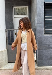 Cappotto di lana marrone chiaro con grandi bottoni, indossato su un gilet di maglia crema e una camicia bianca, abbinato a pantaloni in corduroy beige e una borsa bianca.