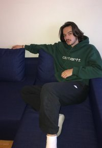 Grøn Carhartt hoodie, sorte sweatpants, sidder på en mørkeblå sofa med to puder, ensartet hvid væg i baggrunden.