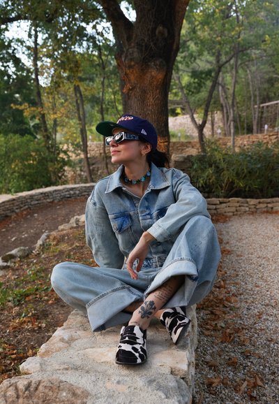 Mono de mezclilla en un tono azul claro, gorra de béisbol, gafas de sol XL y zapatos tipo slip-on con un patrón en blanco y negro. Accesorios incluyen un collar de cuentas.
