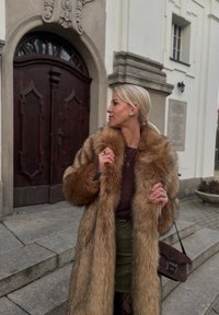 Manteau en fausse fourrure aux tons bruns avec un design long et fluide. Associé à une jupe vert foncé et portant un sac à bandoulière marron foncé.