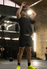 Atleta con maglietta a maniche corte nera e pantaloncini, che esegue un salto. Scarpe da ginnastica neon gialle. Ambiente di palestra industriale con mattoni a vista.