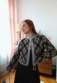 Γυναίκα με κόκκινα μαλλιά, που φοράει γκρι και καφέ καρό ζακέτα και μαύρη φούστα, κρατώντας μια τσάντα, στέκεται σε ένα φωτισμένο ήπια δωμάτιο με ξύλινα έπιπλα.