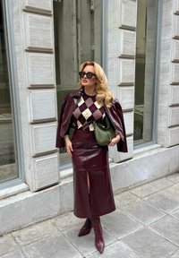 Donna che indossa una giacca e una gonna in pelle color bordeaux, un maglione a rombi, occhiali da sole e tiene una borsa verde, in piedi su un marciapiede urbano.