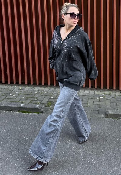 Sudadera negra con cremallera y adornos de piedras brillantes, combinada con vaqueros acampanados gris claro y botas negras de tacón brillante, caminando sobre el pavimento.