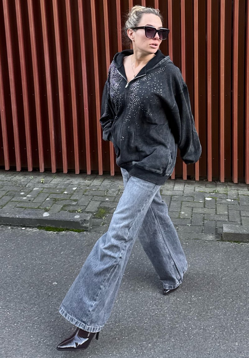 Sweat à capuche noir zippé orné de strass, associé à un jean évasé gris clair et des bottes à talons noires brillantes, marchant sur le trottoir.