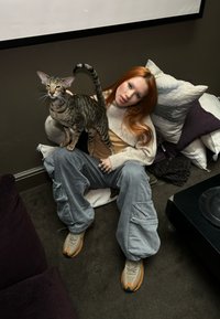 A person wearing a beige knitted sweater and grey cargo trousers, seated with a tabby cat on their lap, surrounded by cushions on a dark floor.