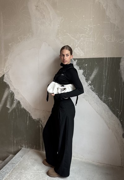 Mujer con atuendo negro sosteniendo una bolsa blanca, de pie de lado contra una pared gris con patrones abstractos en blanco y gris oscuro.
