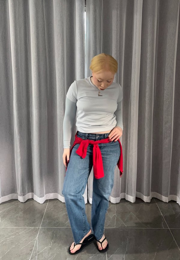 Giovane donna con capelli biondi che indossa una maglietta a maniche lunghe grigio chiaro, jeans blu, maglione rosso legato in vita e infradito nere, in piedi all'interno.