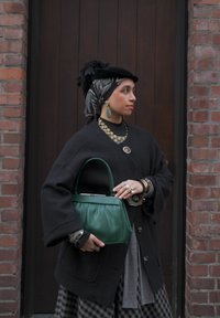 Femme portant un long manteau sombre et un hijab à motifs, tenant un sac à main vert, debout devant une porte en bois encadrée par des murs en briques.