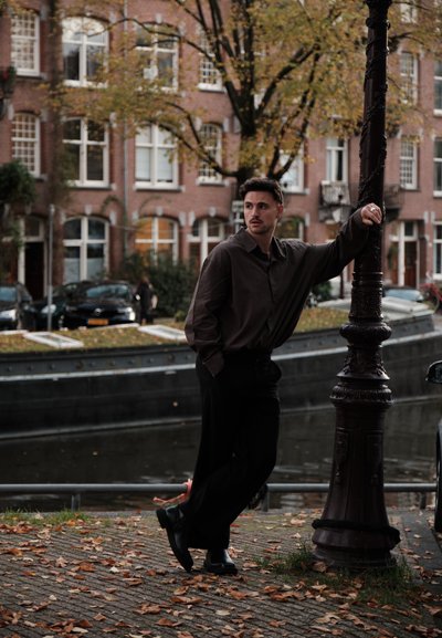 Camisa de botones marrón oscuro, pantalones negros y zapatos de cuero. Apoyado en una farola decorativa junto a un canal en un entorno urbano con hojas de otoño.