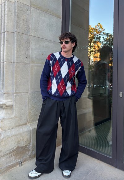 Suéter de punto con base en azul marino y patrón argyle en rojo, blanco y azul. Combinado con pantalones negros sueltos y zapatos con estampado.