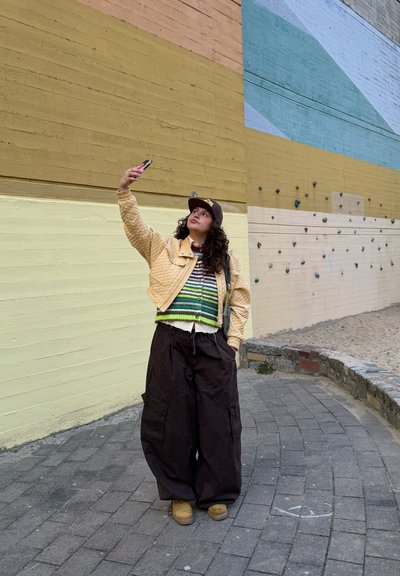 Persona con chaqueta acolchada beige, suéter a rayas y gorra se toma una selfie frente a un muro de escalada multicolor al aire libre.