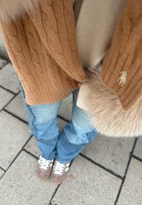 Άτομο που φοράει ανοιχτό μπλε τζιν, μπεζ αθλητικά παπούτσια, κρεμ πουλόβερ με χνουδωτά μανίκια και καφέ πλεκτή ζακέτα με σχέδιο καλώδιου, στέκεται πάνω σε πλακόστρωτο πεζοδρόμιο.