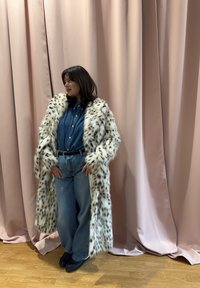 Femme portant un long manteau en fausse fourrure blanche à taches noires, une chemise en jean et un jean large, debout sur un sol en bois devant des rideaux roses.
