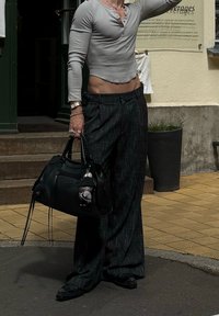 Siva dolga rokavna henley majica, ohlapne črne hlače s vzorcem in črna torbica z obeskom. Teren je tlakovan, ozadje pa je zeleno.